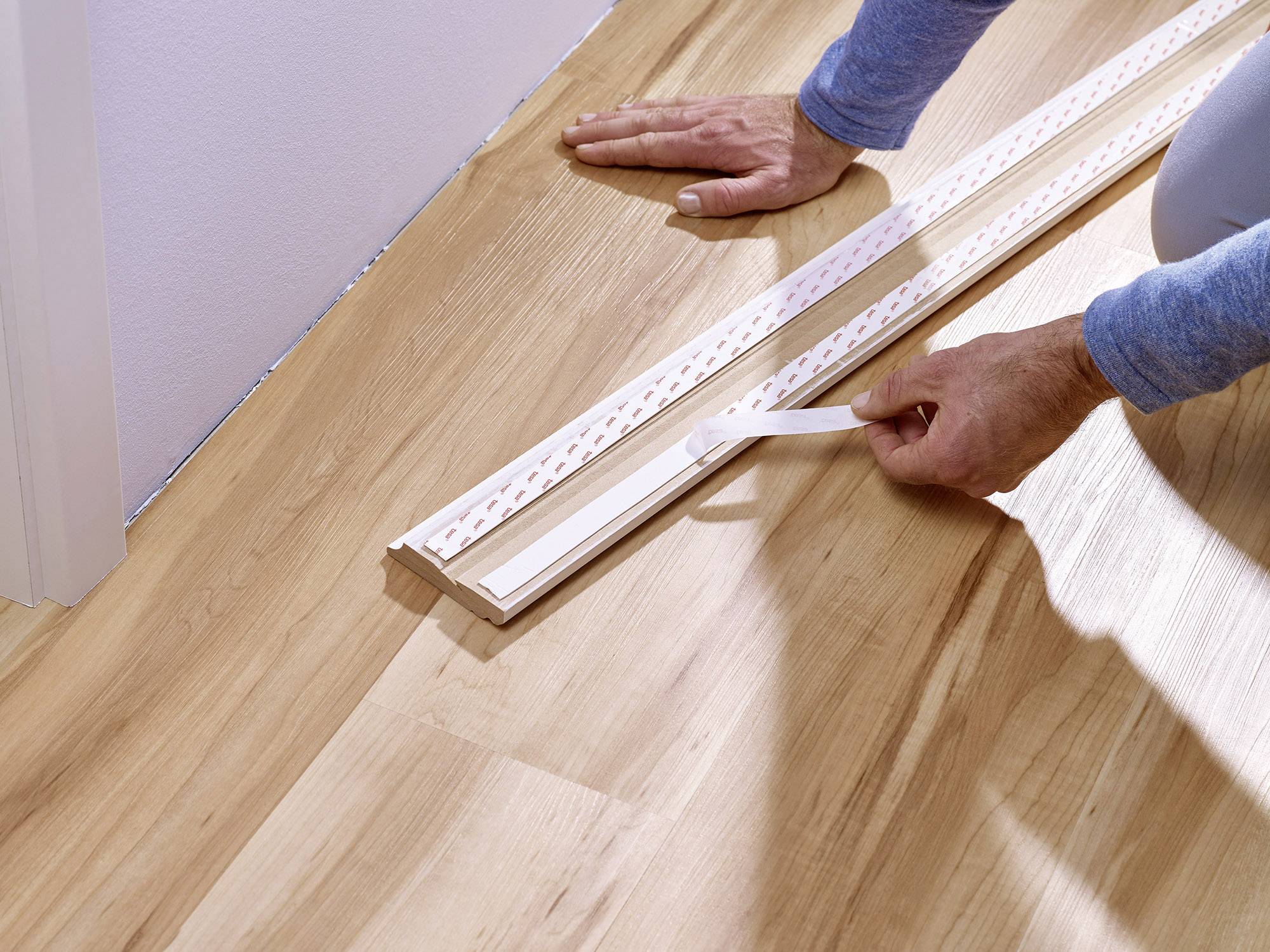 Une personne installe une barre de seuil sur un plancher en bois près d'un mur. Les mains tiennent l'outil et la barre.