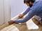 Un homme installe soigneusement une plinthe blanche sur un plancher en bois le long du mur.