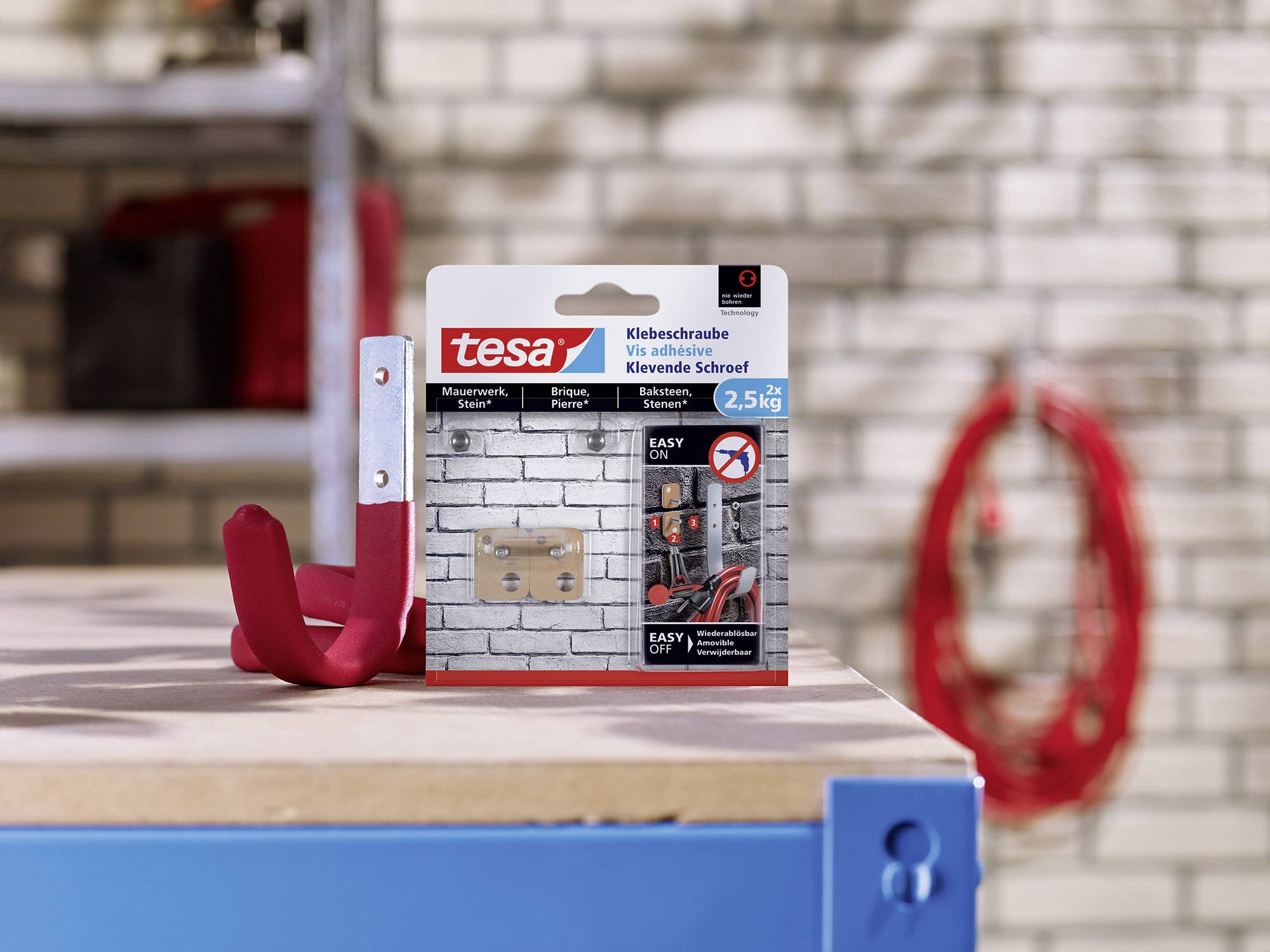 Boîte de vis adhésives tesa posée sur une table devant un mur de briques. Ces vis sont adaptées aux surfaces solides et peuvent supporter jusqu'à 2,5 kg.