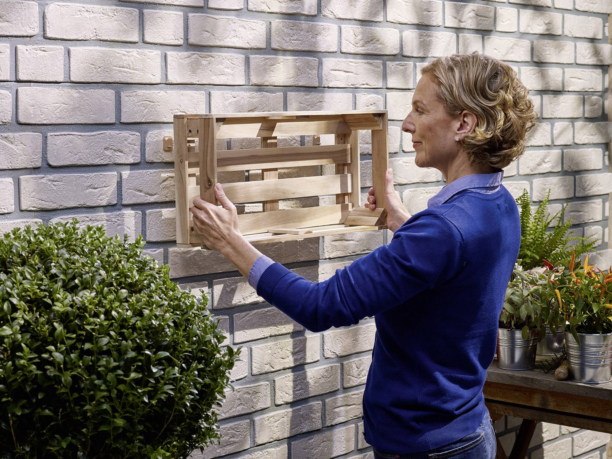 Une personne installe une étagère en bois sur un mur de briques dans le jardin. Les plantes à proximité créent une atmosphère naturelle.
