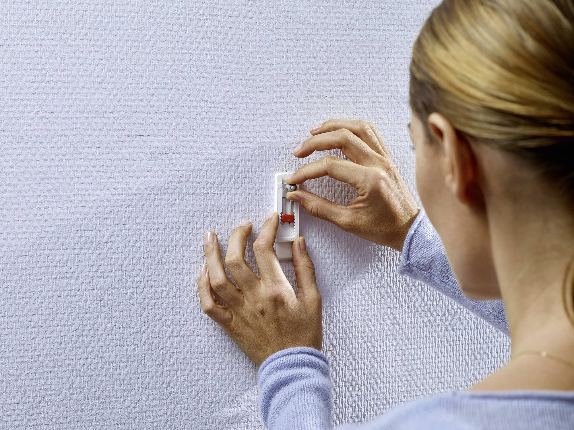 Une personne fixe un petit objet sur un mur texturé en le poussant. Les mains sont au centre de l'image.
