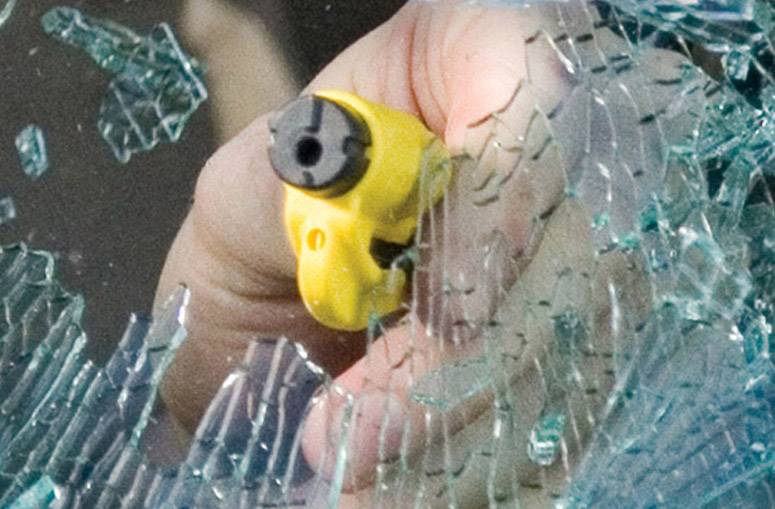 Une main tient un outil jaune et le pousse à travers du verre brisé pour enfoncer une fenêtre. Scène de sécurité ou de situation d'urgence.