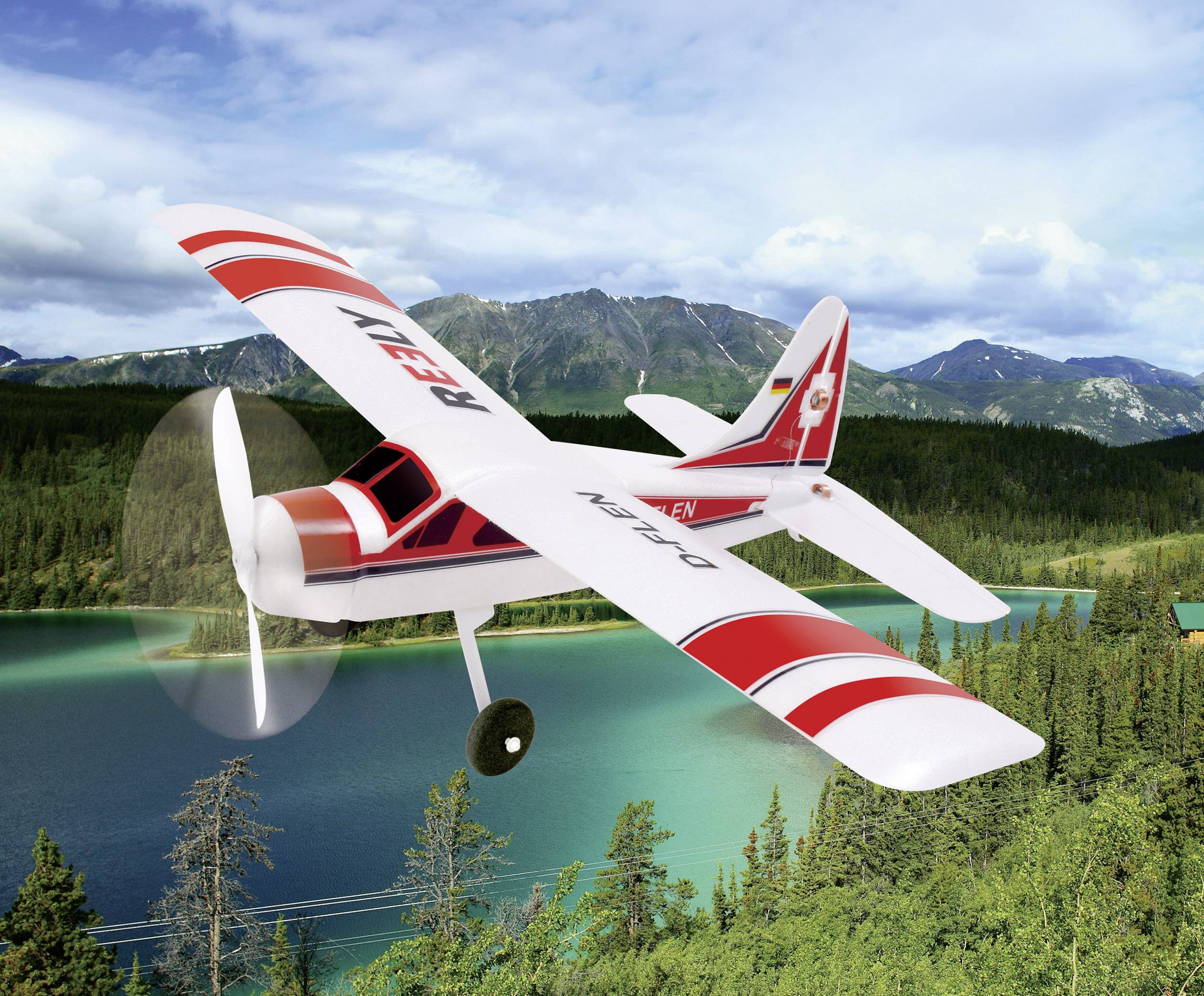 Un avion-modèle rouge et blanc survole un lac turquoise, entouré de collines boisées et de montagnes en arrière-plan.