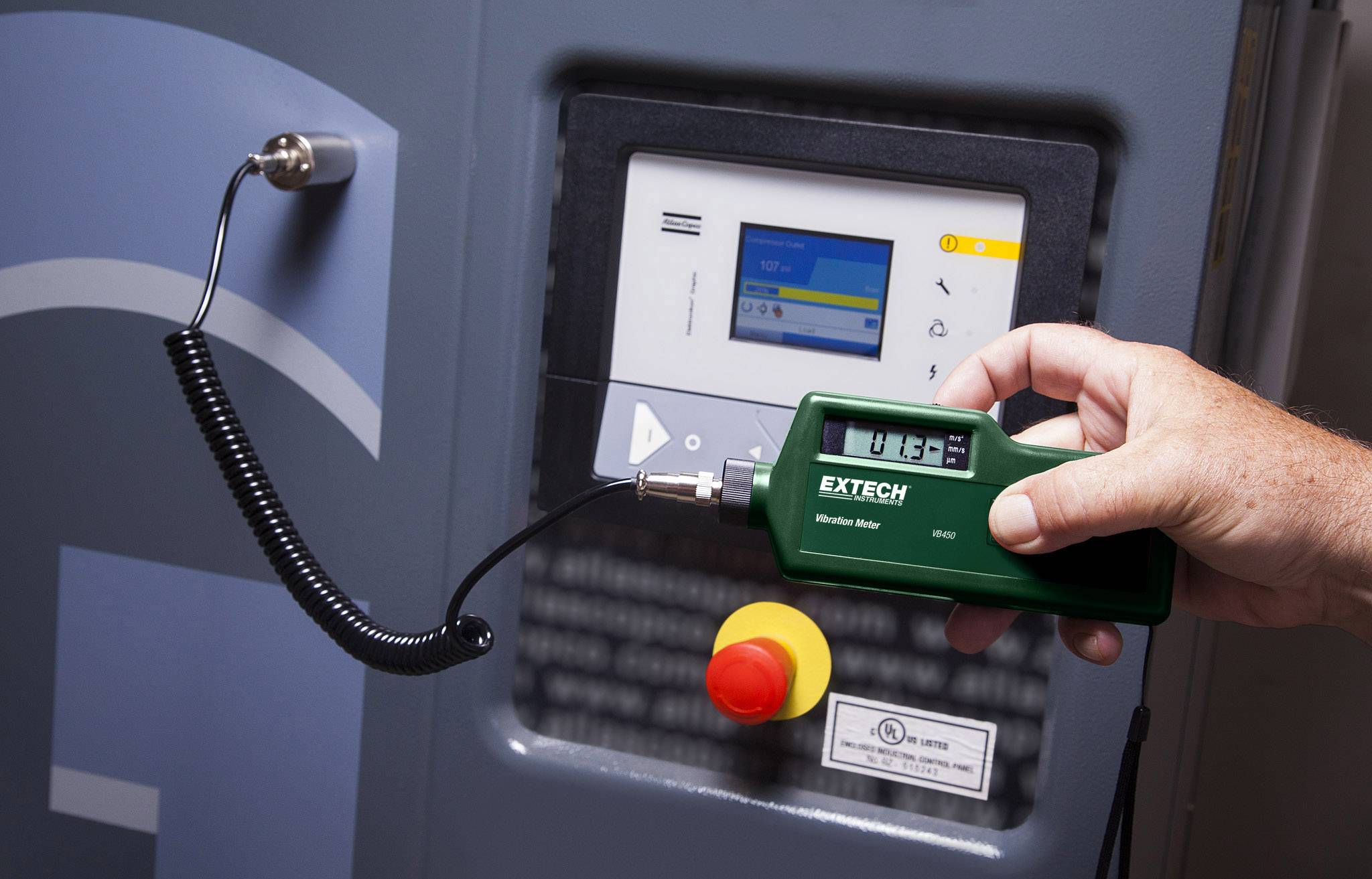 Une personne tient un appareil de mesure vert devant un panneau de commande électronique avec un écran. Le panneau de commande possède plusieurs touches colorées.