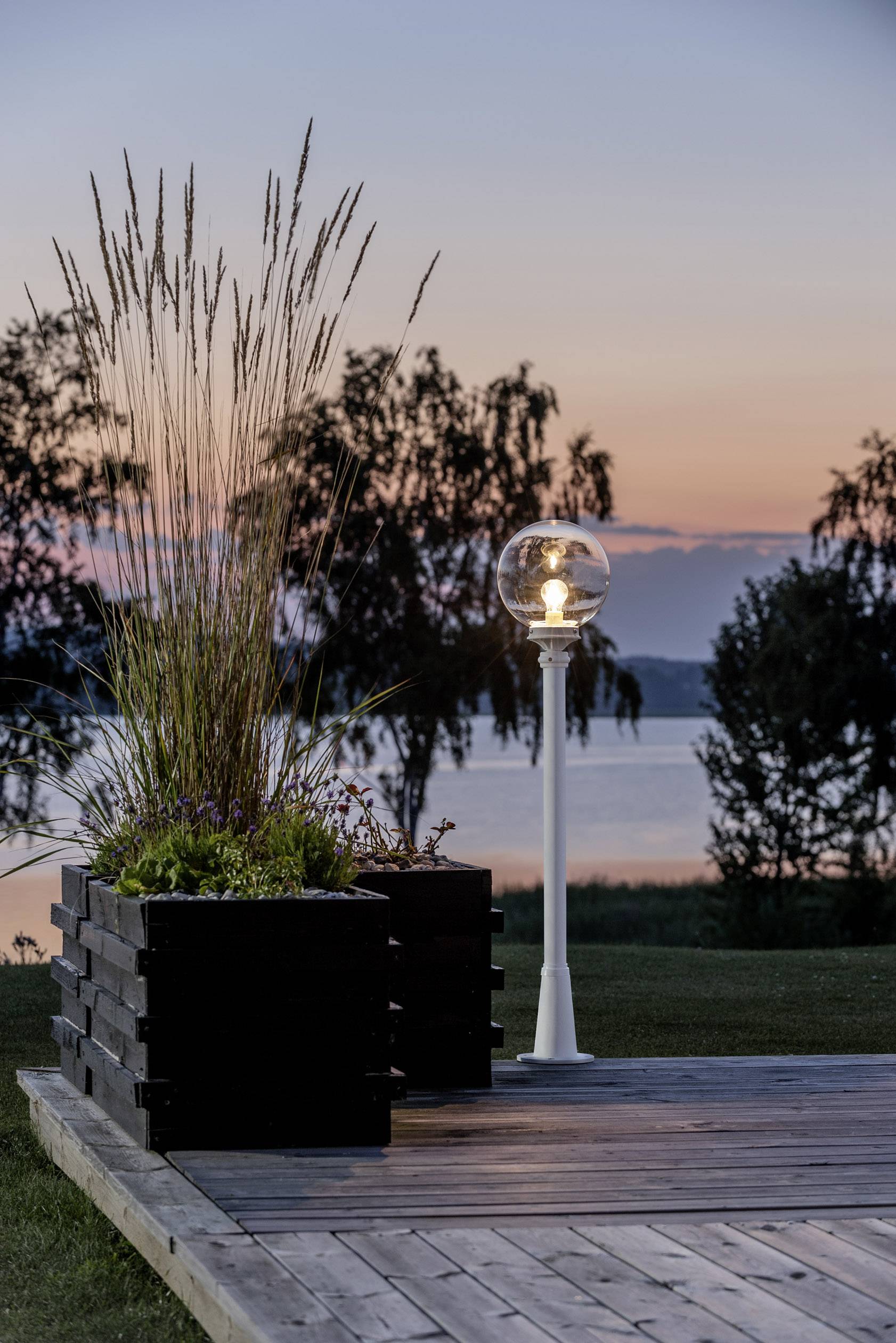 Terrasse en bois avec des jardinières et une lampe ronde au premier plan, lac et arbres en arrière-plan au coucher du soleil.