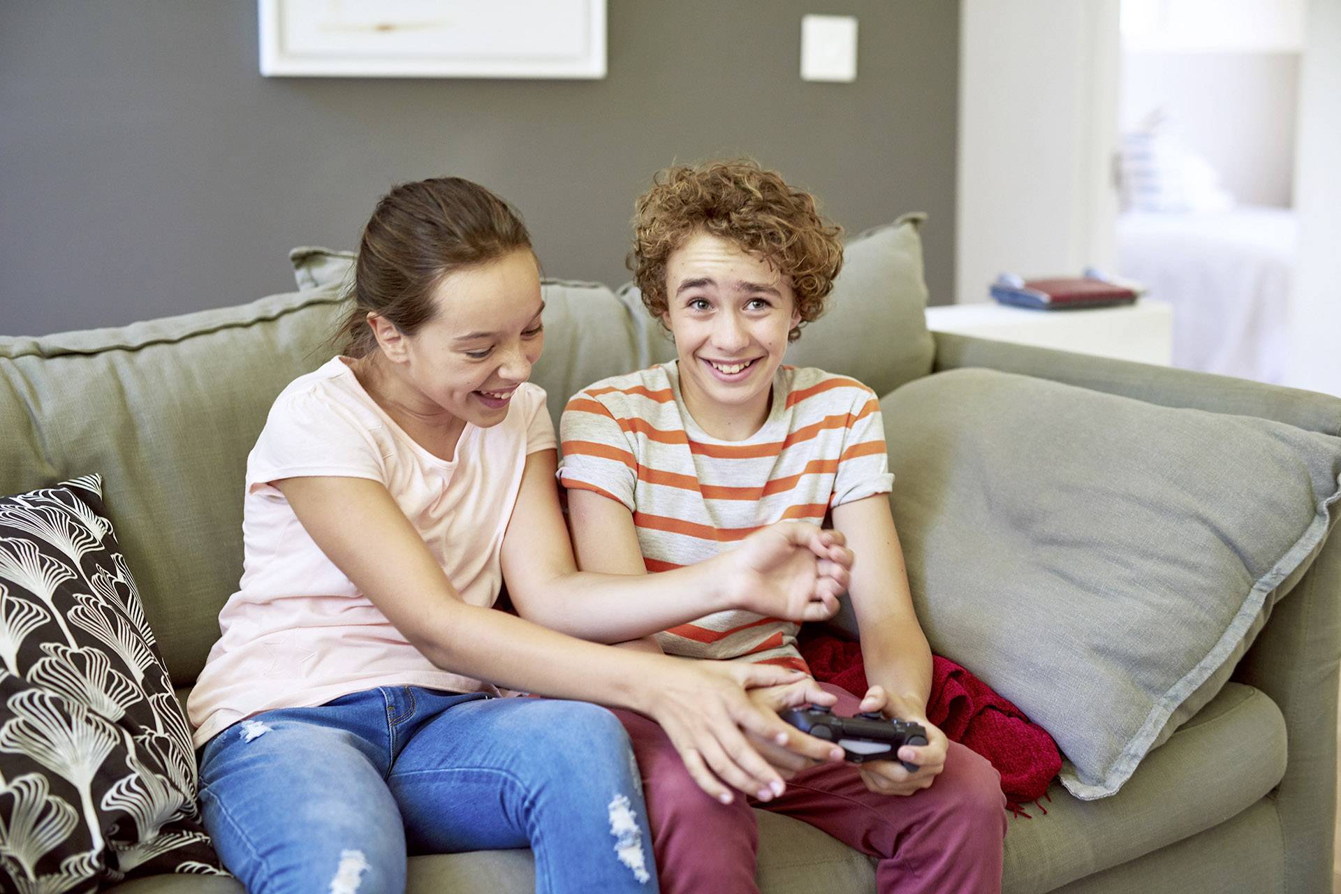 Deux enfants sont assis sur un canapé, riant et jouant à des jeux vidéo avec une manette à la main.