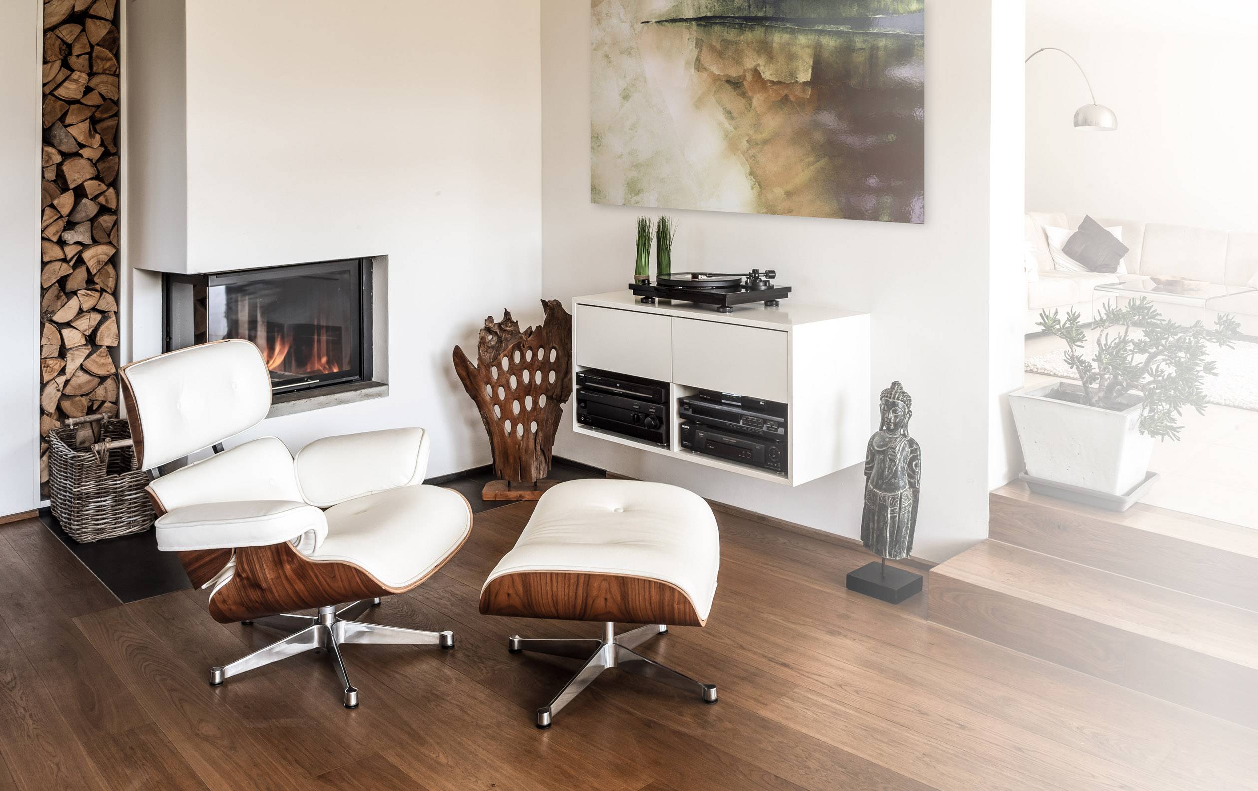 Salon avec un fauteuil blanc et un tabouret, cheminée, pile de bois, art moderne sur le mur et une chaîne stéréo sur une étagère murale.