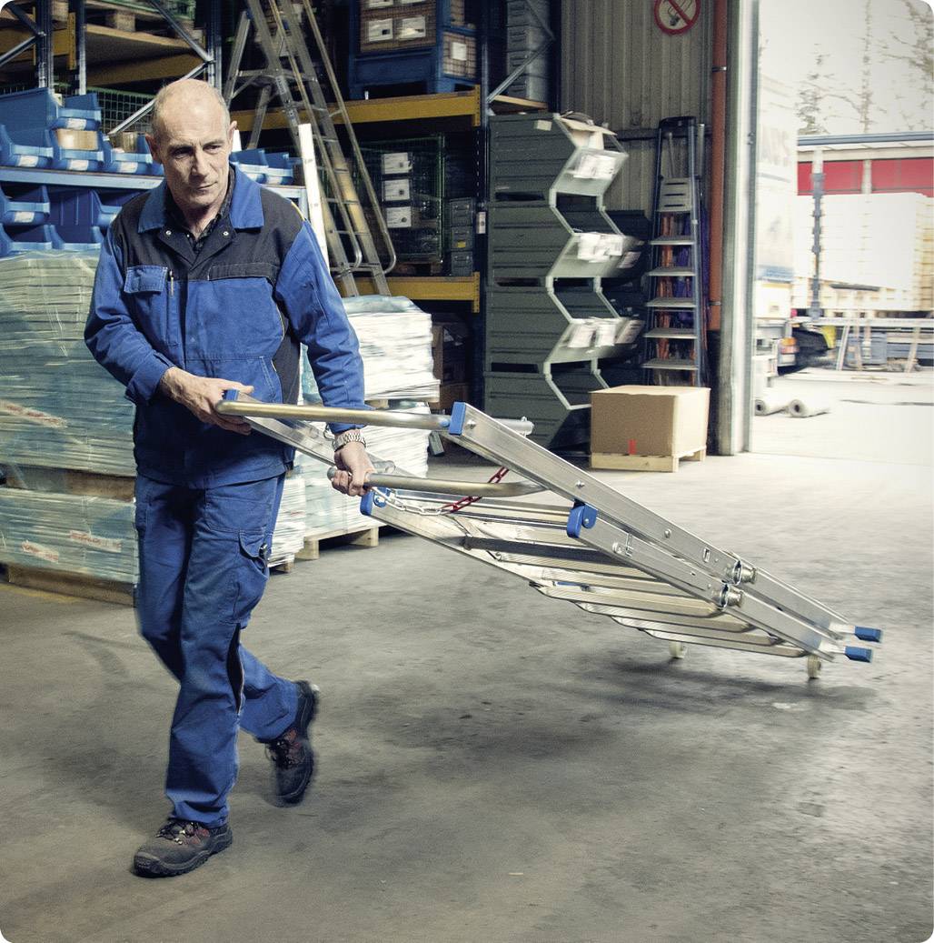 Un homme en vêtements de travail porte une échelle pliée à travers un entrepôt. Des étagères et des cartons sont visibles en arrière-plan.