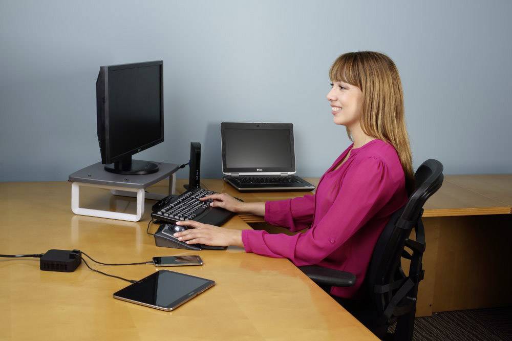 Une femme est assise à un bureau avec deux écrans, un ordinateur portable, une tablette et un smartphone, souriant en travaillant.