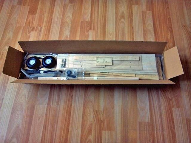 Carton contenant des pièces de meubles en bois démontées et des roulettes sur un plancher en bois, prêt à être assemblé.