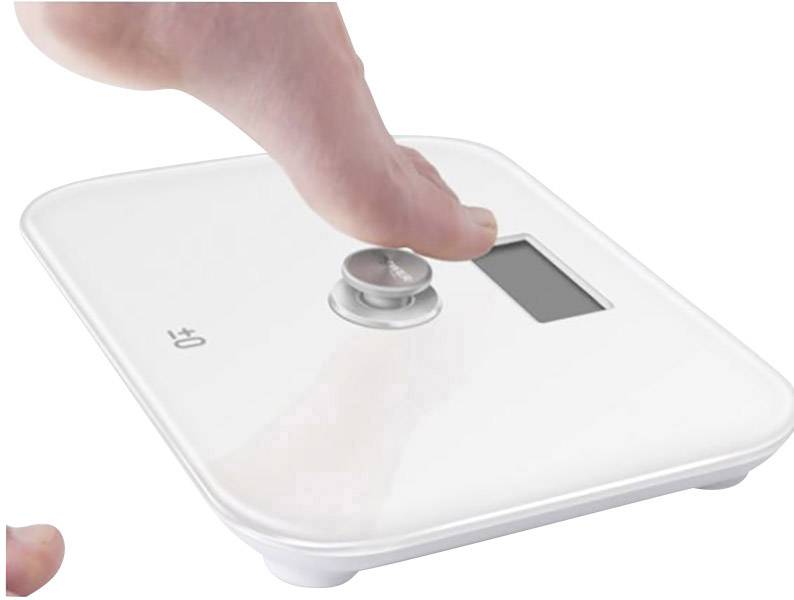 Un pied nu actionne un pèse-personne numérique à boîtier blanc avec un écran affichant le poids en kilogrammes.