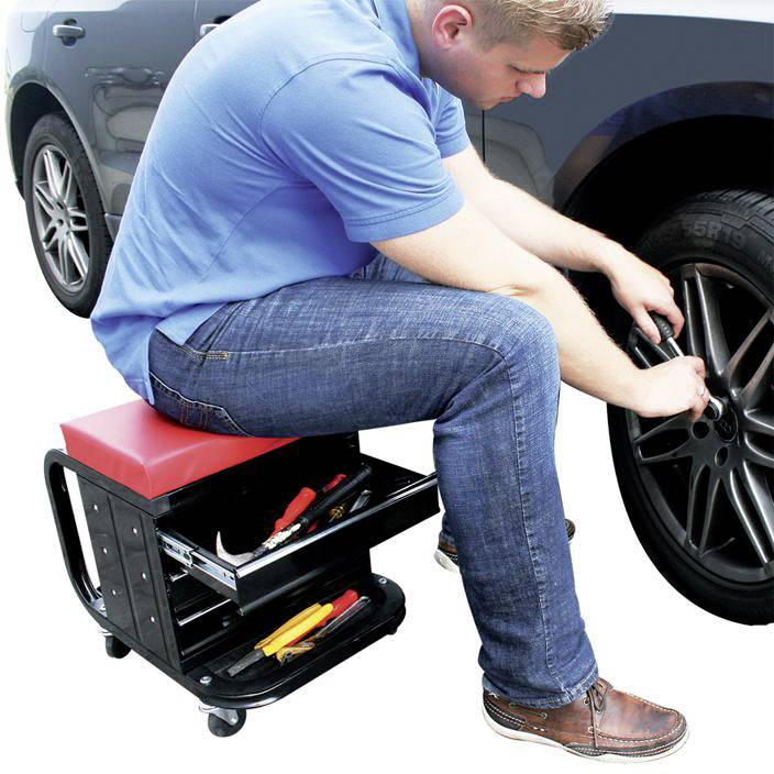 Un homme change un pneu dans un atelier. Il est assis sur un tabouret mobile avec un espace de rangement pour les outils et saisit la roue.