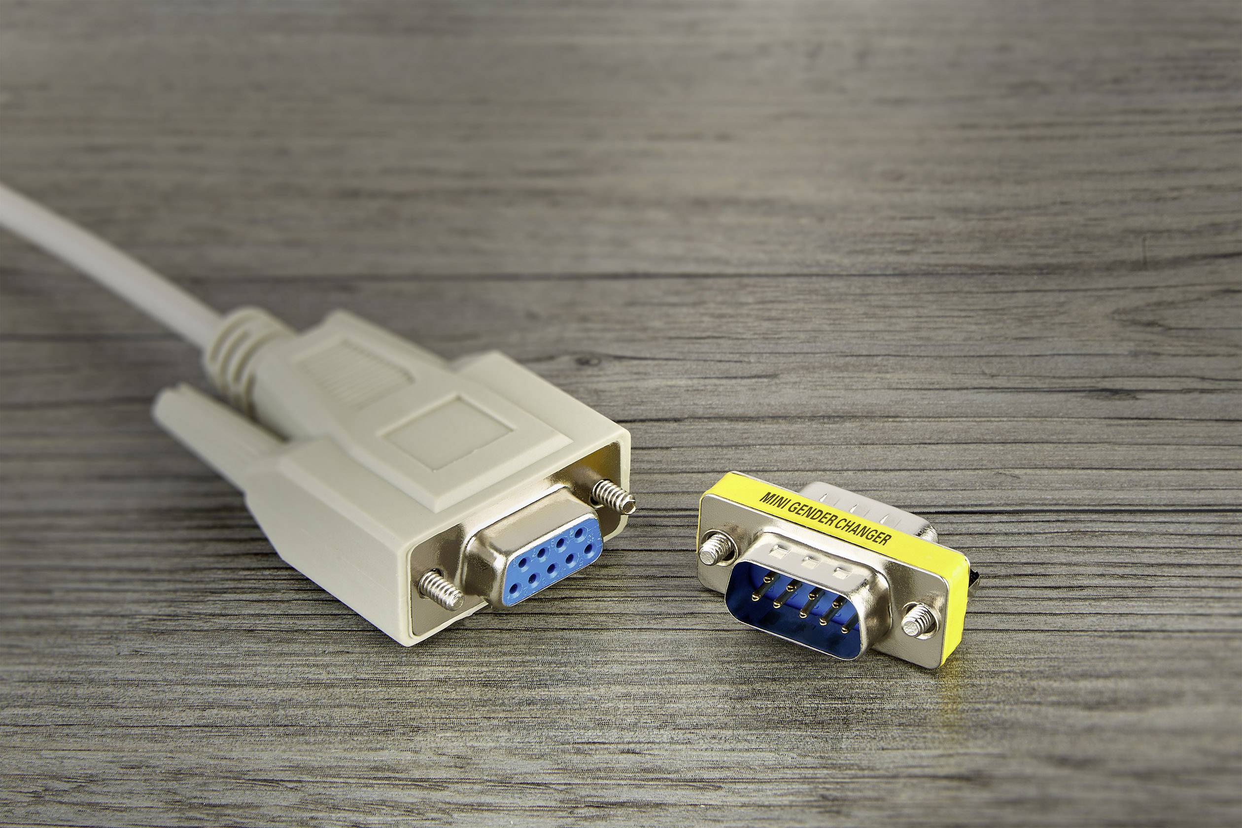 Un connecteur VGA et un adaptateur de genre VGA sur une surface en bois. La fiche a des broches bleues, l'adaptateur est étiqueté en jaune.