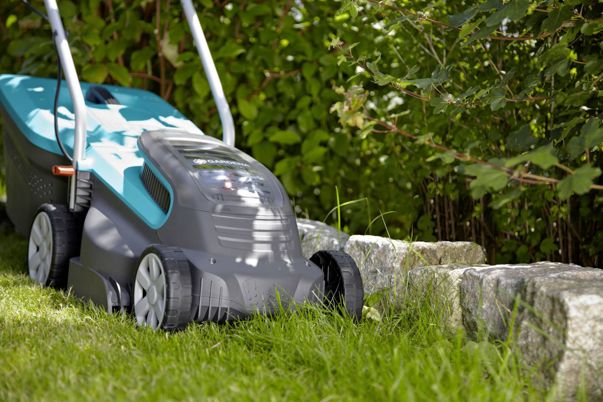 Tondeuse électrique tondant l'herbe à côté d'un massif de pierres et d'arbustes. Idéale pour les petits et moyens jardins.