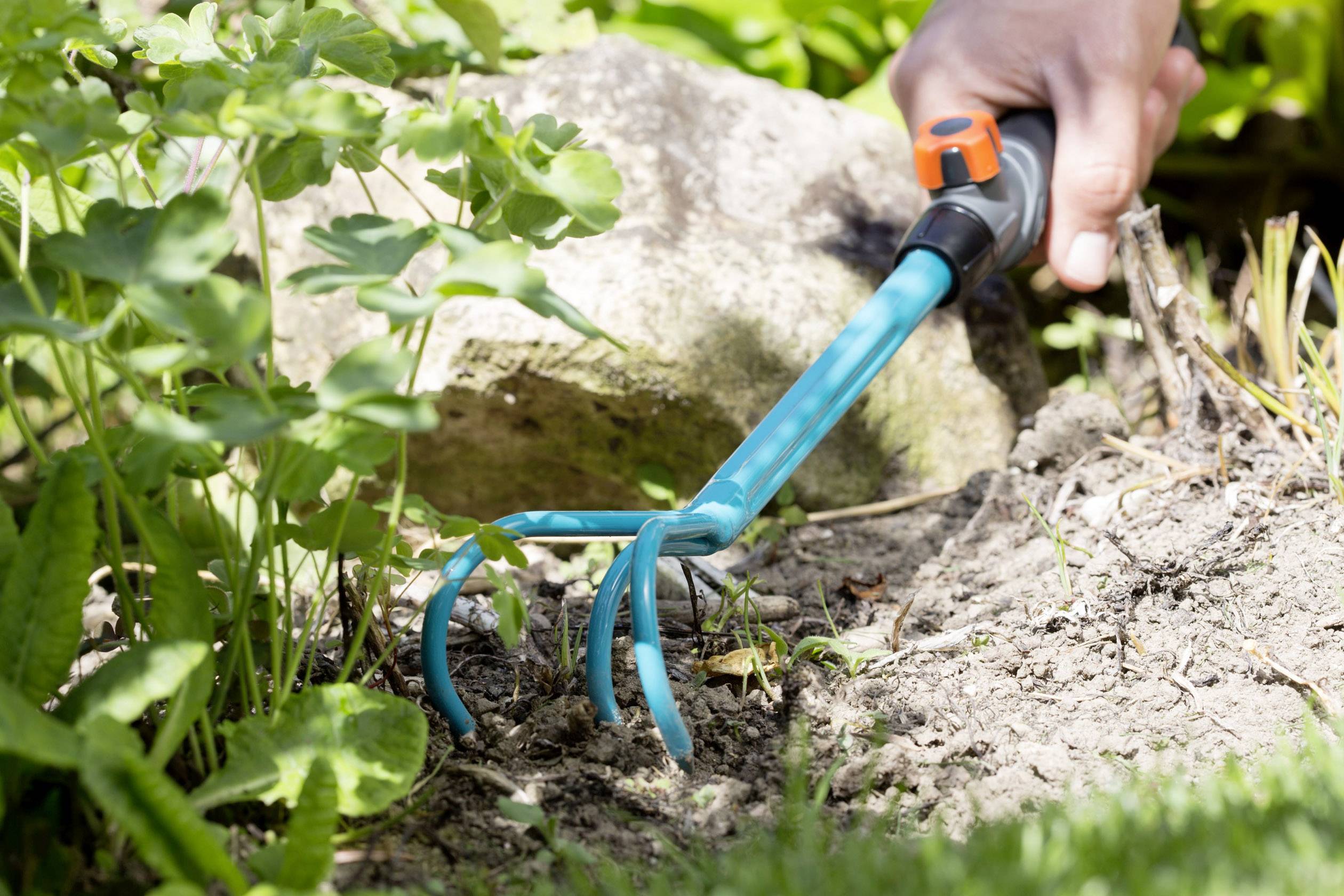 Une main tient une houe de jardin et travaille la terre dans un jardin. Des plantes et des pierres sont visibles en arrière-plan.