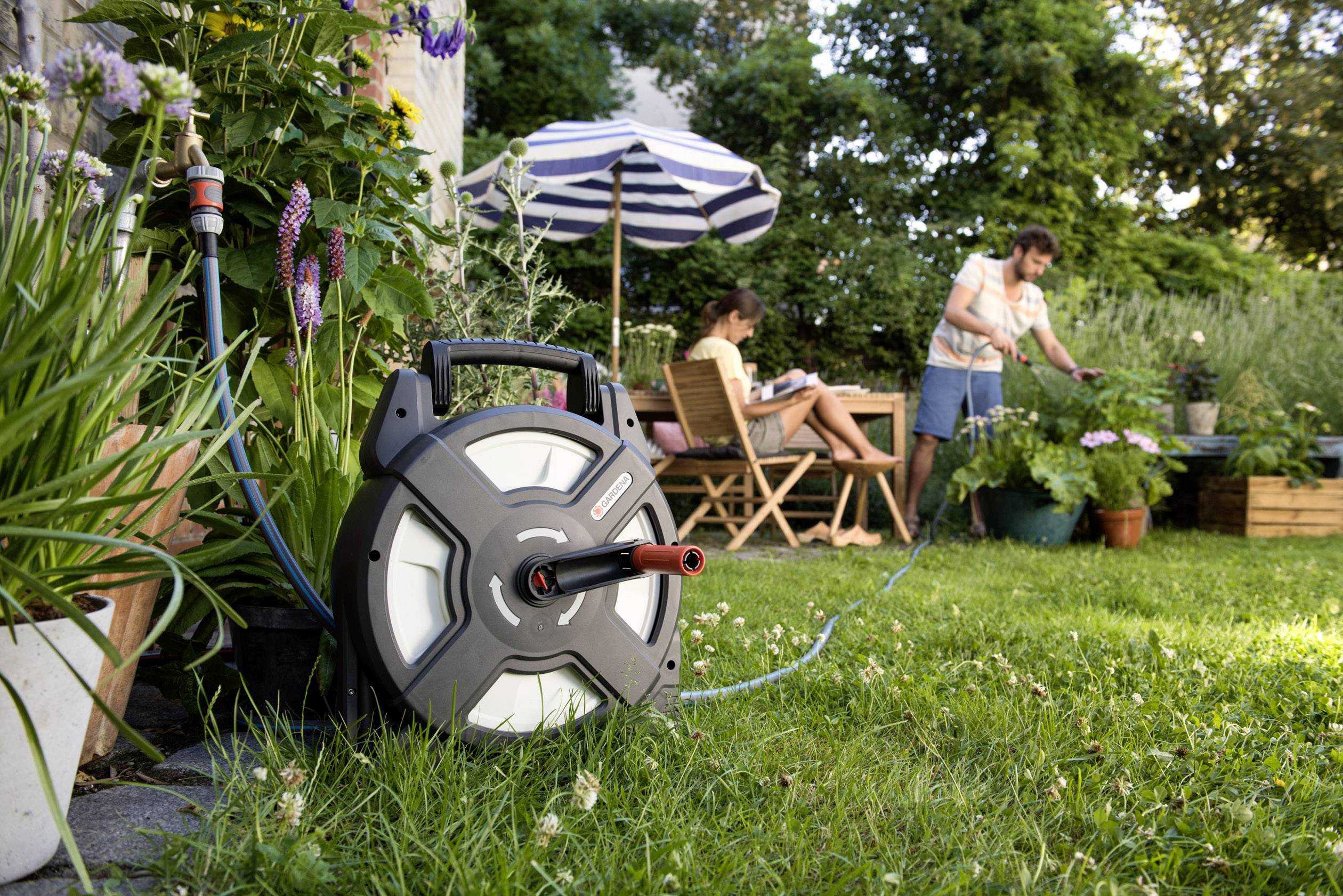 Scène de jardin montrant un homme arrosant des plantes pendant qu'une femme lit assise sur une chaise ; au premier plan, un tuyau d'arrosage enroulé sur un dévidoir.