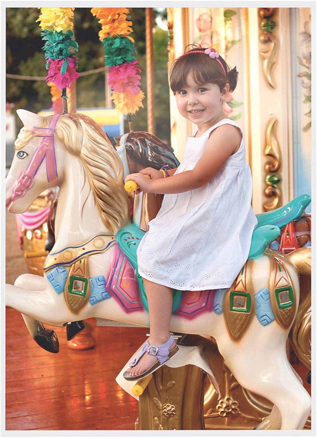 Un enfant en robe blanche sourit assis sur un cheval de carrousel. Des décorations colorées sont visibles en arrière-plan.