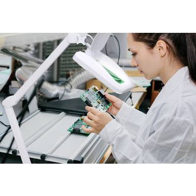 Une ingénieure en blouse blanche inspecte une carte électronique sous une loupe dans un laboratoire.