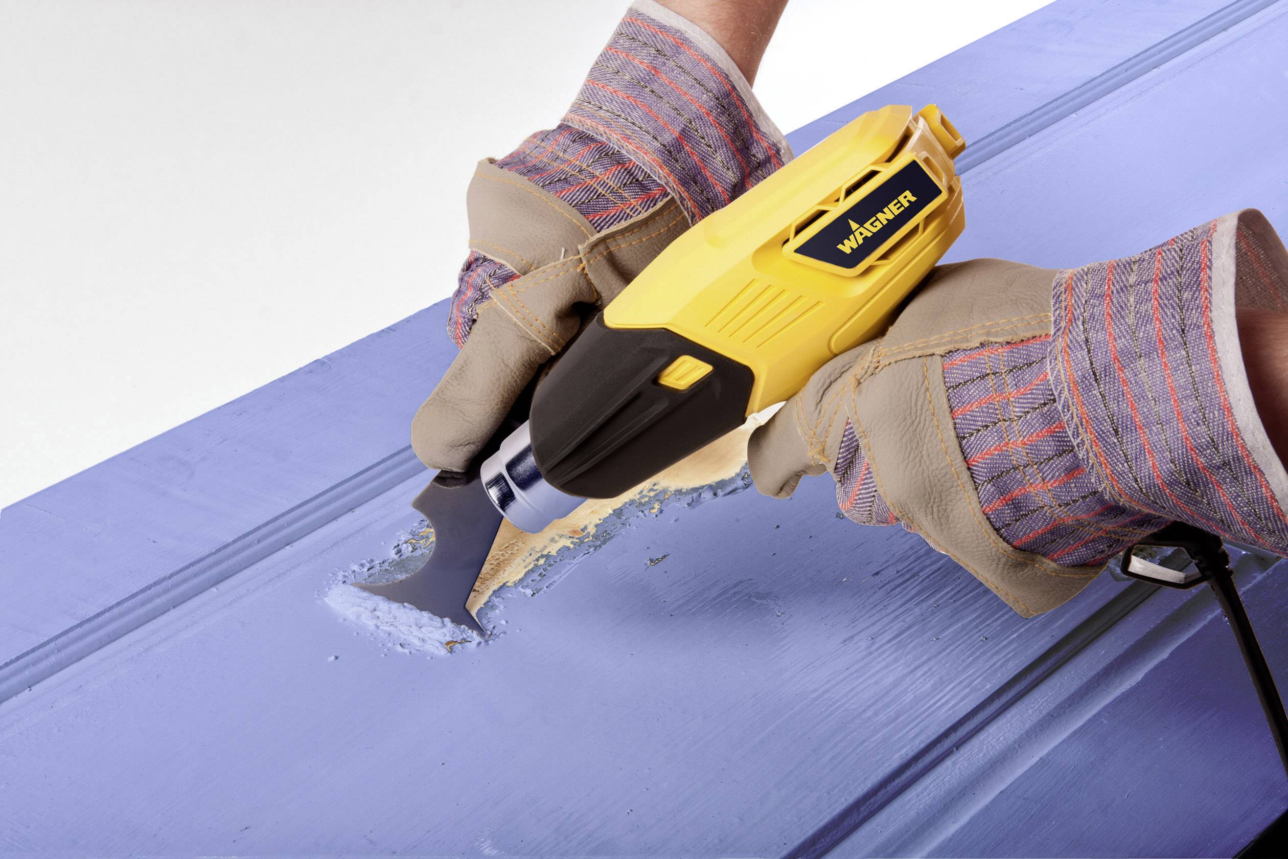Une personne portant des gants de travail enlève de la peinture d'une surface en bois à l'aide d'un décapeur électrique jaune.