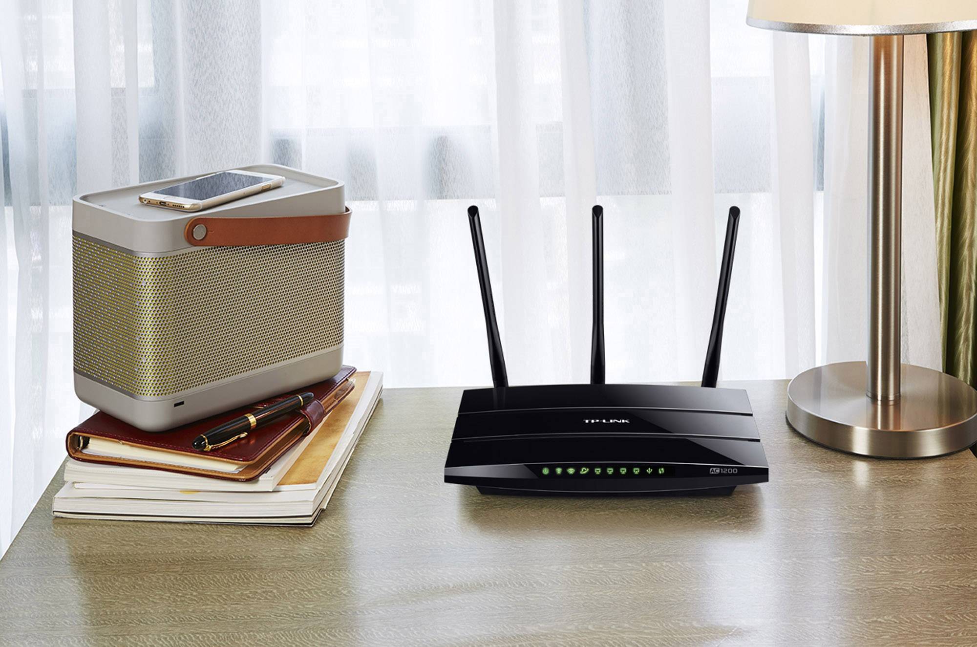 Un routeur noir à trois antennes posé sur un bureau à côté de livres empilés et d'un haut-parleur, avec des rideaux en arrière-plan.