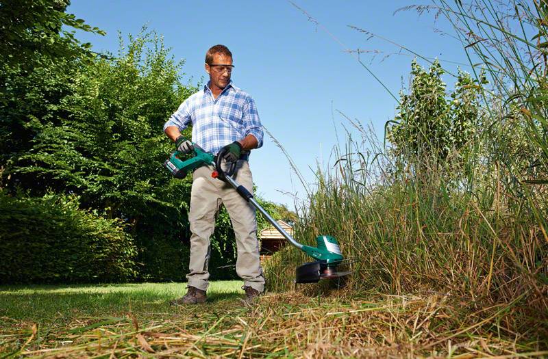 Un homme portant des lunettes de sécurité et des gants taille l'herbe et les buissons dans le jardin avec un outil électrique par temps ensoleillé.