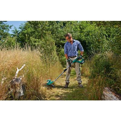 Un homme tond l'herbe avec un coupe-bordures électrique dans un jardin naturel, entouré de buissons et d'arbres élevés.