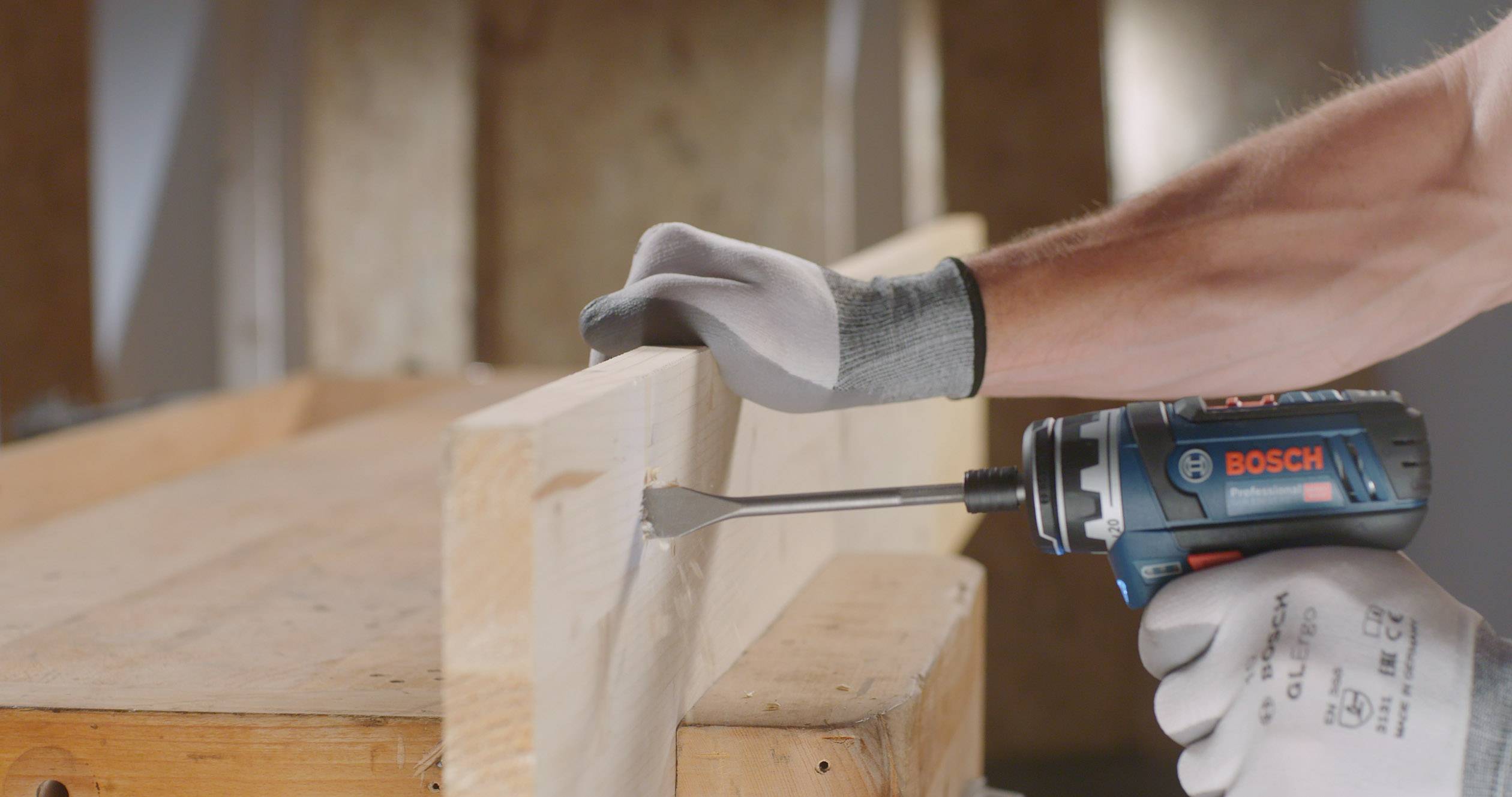 Une personne utilise une perceuse à percussion Bosch bleue, alimentée par batterie, pour percer un trou dans une planche en bois. Porter des gants.