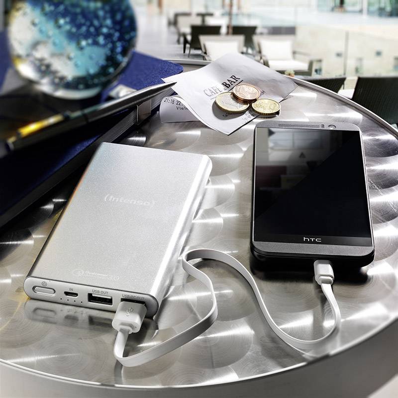 Une batterie externe charge un smartphone sur une table ronde argentée. Des documents et des pièces de monnaie sont visibles en arrière-plan.