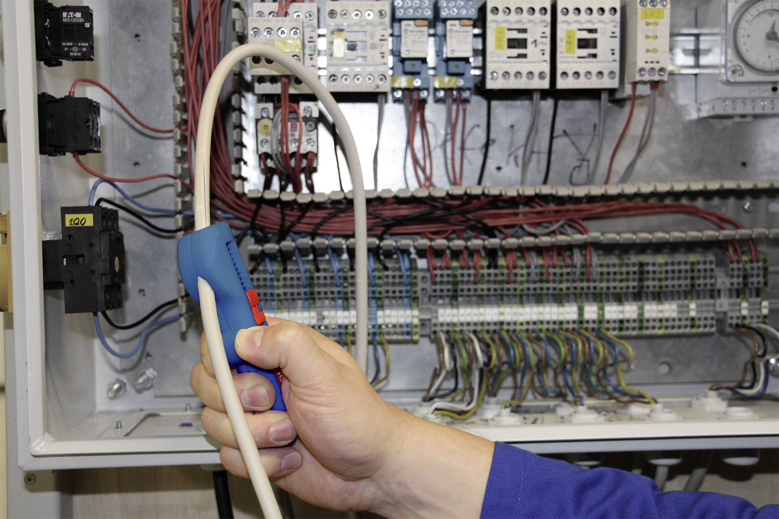 Une main tient un appareil de test devant un armoire électrique ouverte remplie de câbles et de composants électriques, probablement pour une opération de maintenance.