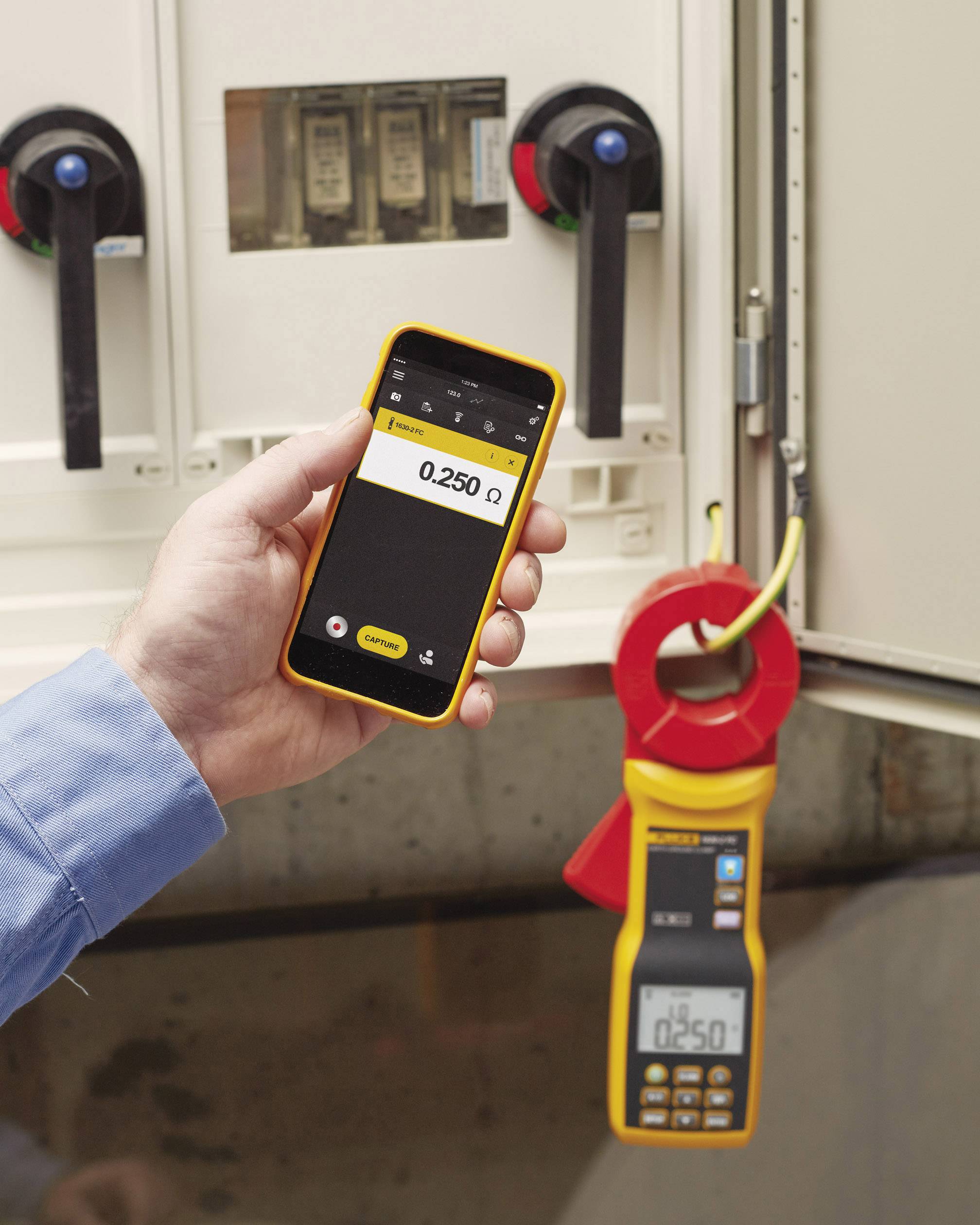 Une personne tient un appareil mobile affichant 0,250 ohm. Un appareil de type multimètre est connecté à un armoire électrique.