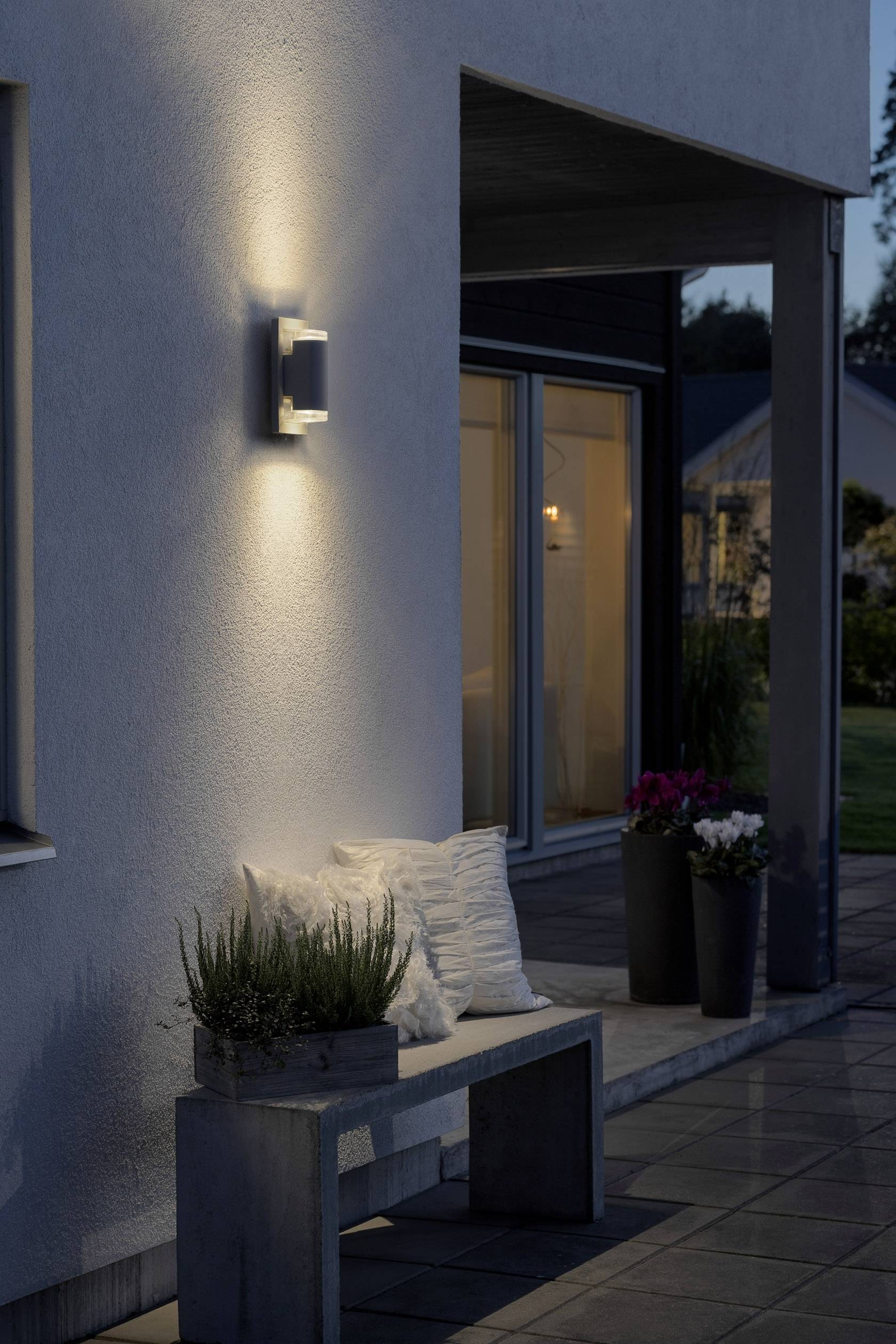 Vue extérieure d'une terrasse moderne au crépuscule avec un applique murale éclairée, des plantes sur un banc en béton et des fenêtres en arrière-plan.