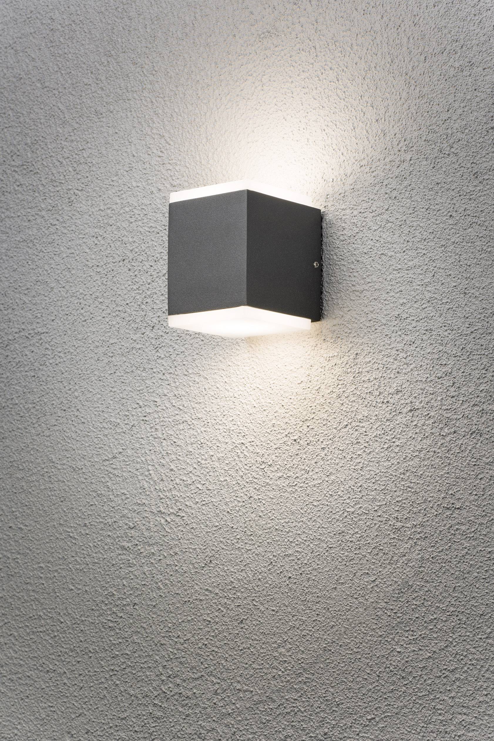 Un luminaire extérieur moderne en noir est fixé sur un mur gris et projette une douce lumière vers le haut et vers le bas.