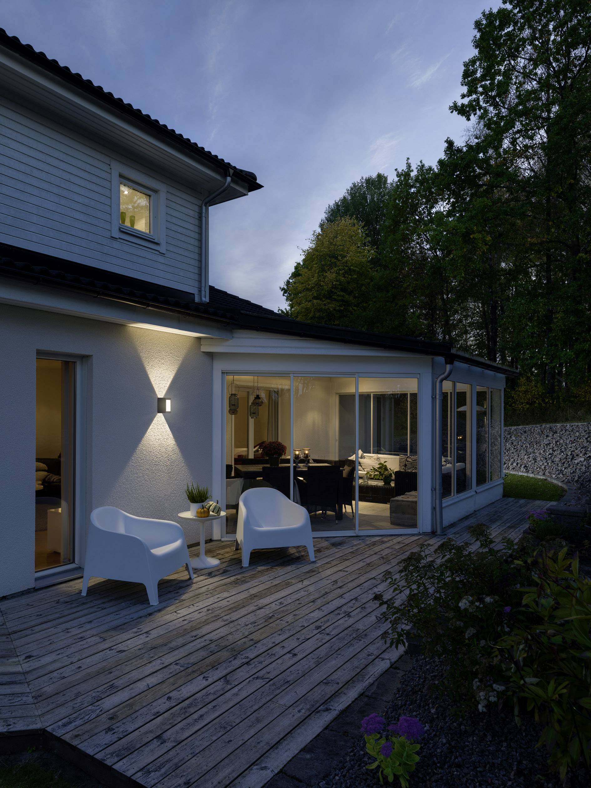 Véranda moderne attenante à une maison individuelle, entourée d'arbres, avec un éclairage du soir. Deux chaises blanches sur la terrasse en bois.