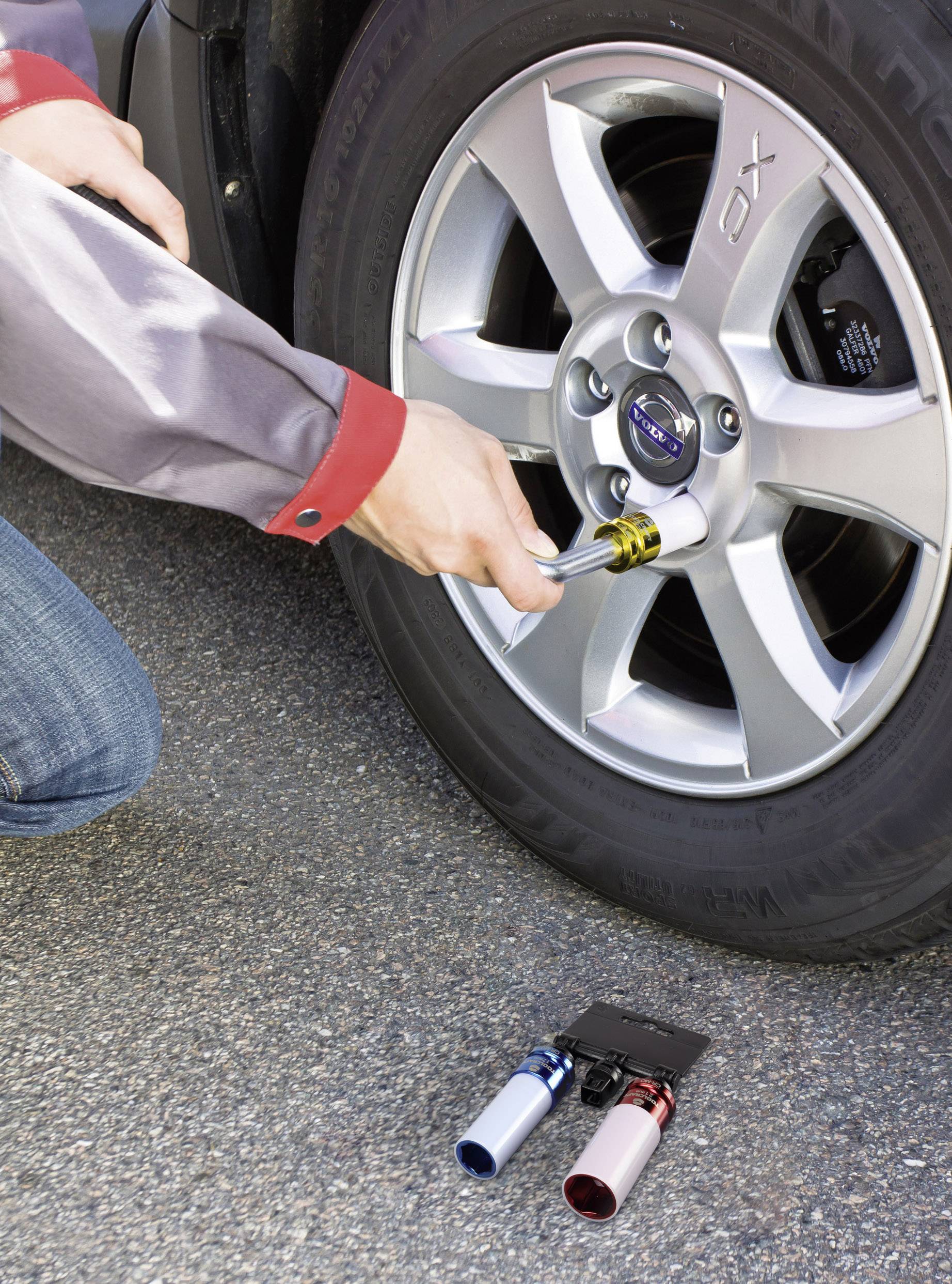 Une personne vérifie la pression d'air dans un pneu de voiture à l'aide d'un manomètre. Deux adaptateurs de valve de pneu sont posés à côté sur le sol.