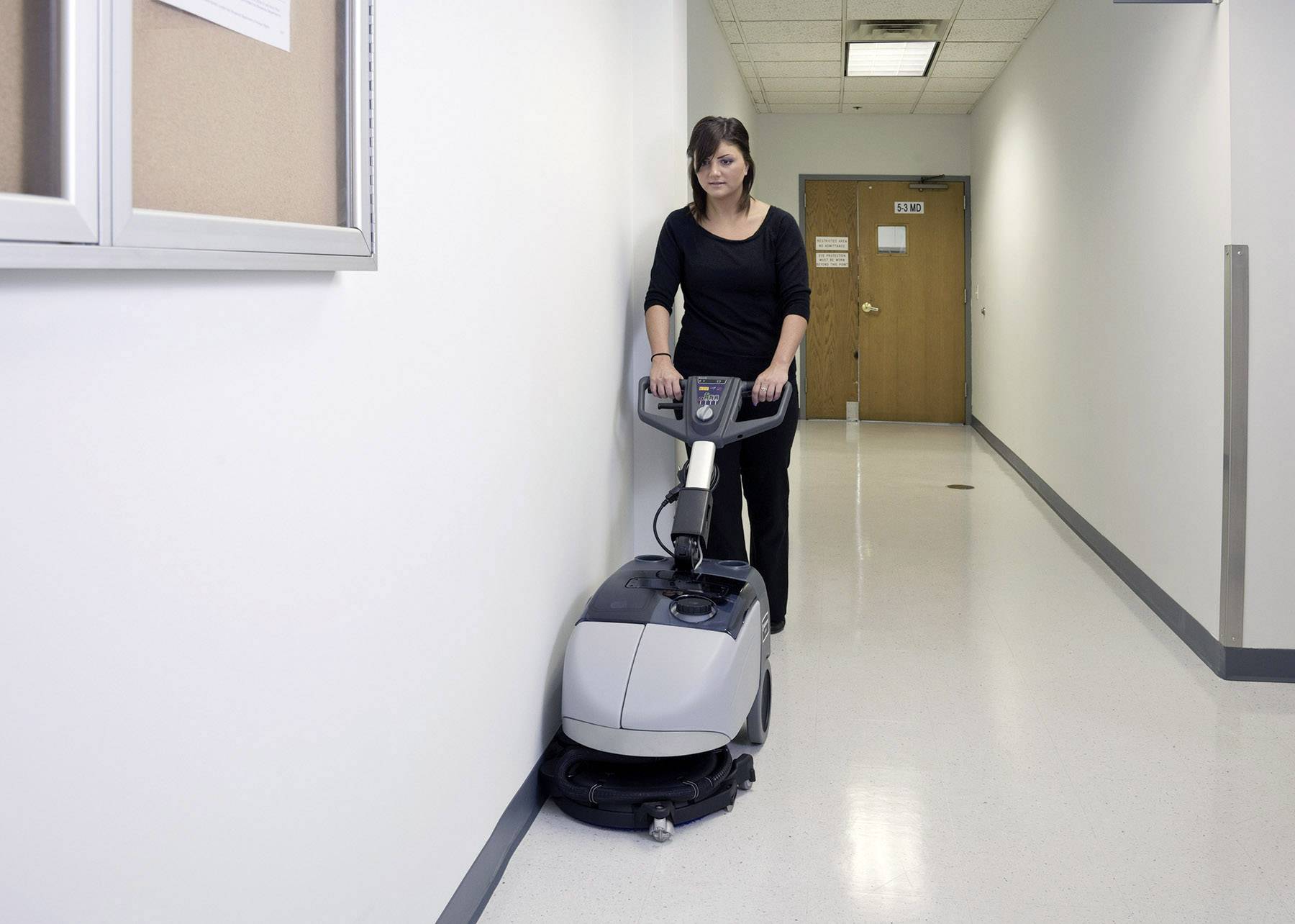Une femme utilise une machine de nettoyage de sol grise dans un couloir de bureau lumineux.