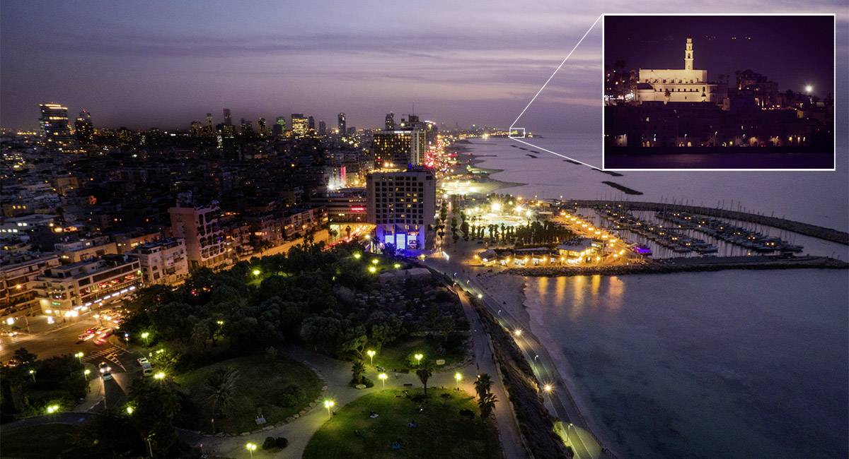Vue sur une ville côtière illuminée la nuit, avec un accent sur un bâtiment éclairé sur une colline en arrière-plan.