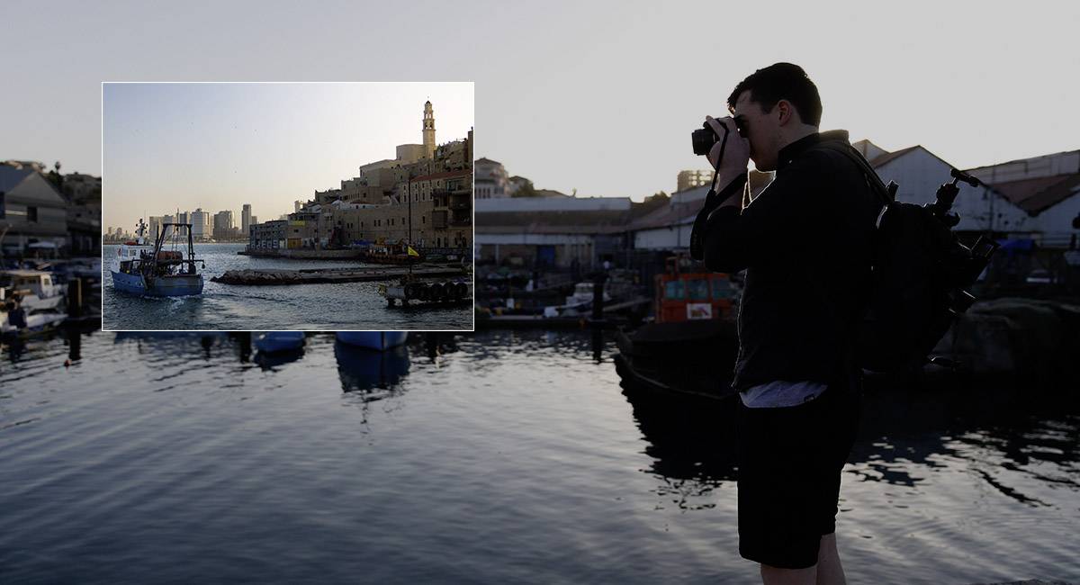 Un photographe se tient sur le port et photographie la côte au coucher du soleil. Des bâtiments et des bateaux sont visibles en arrière-plan.