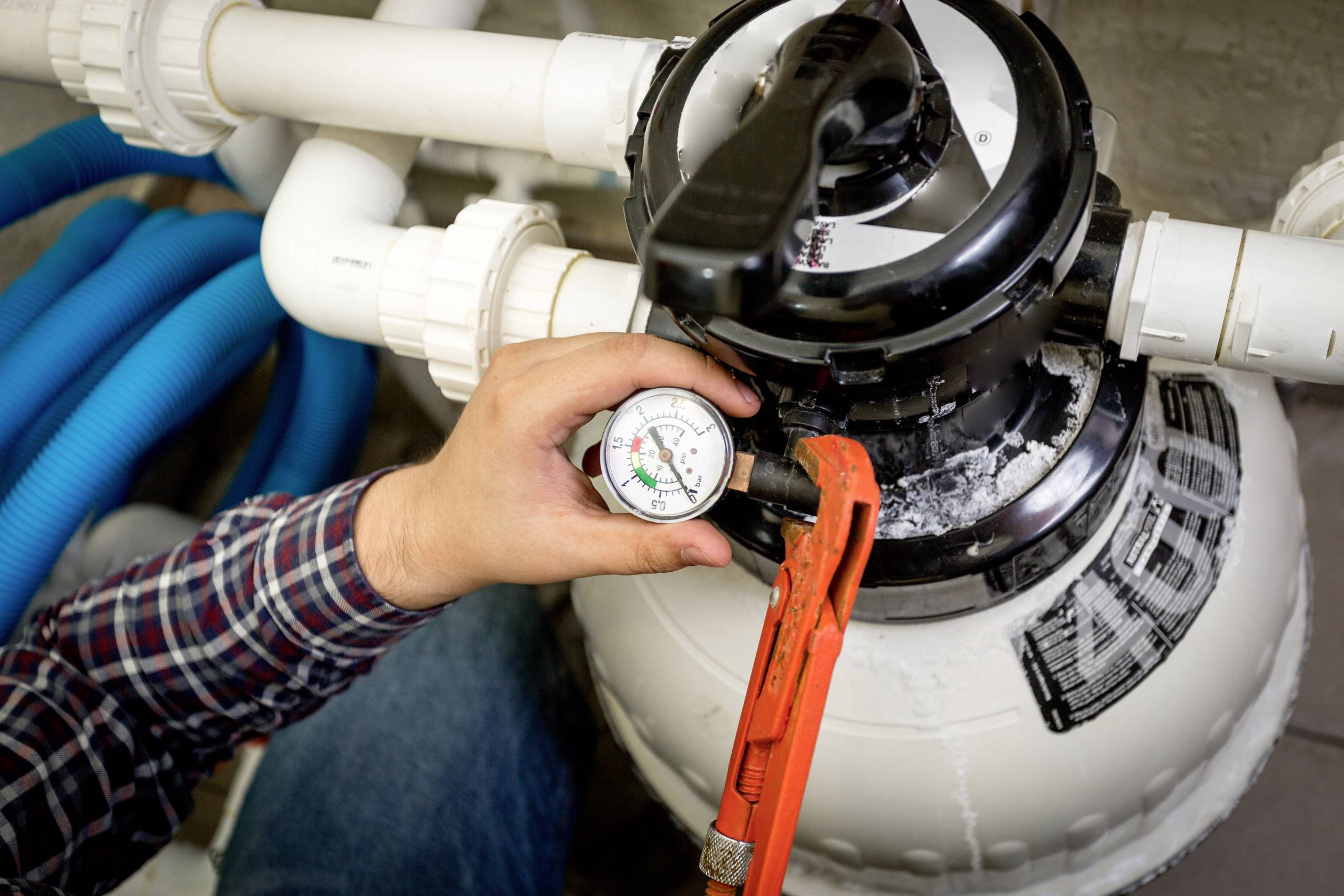 Une personne ajuste le manomètre d'un système de filtration de piscine, une pince à la main. Des tuyaux sont visibles en arrière-plan.