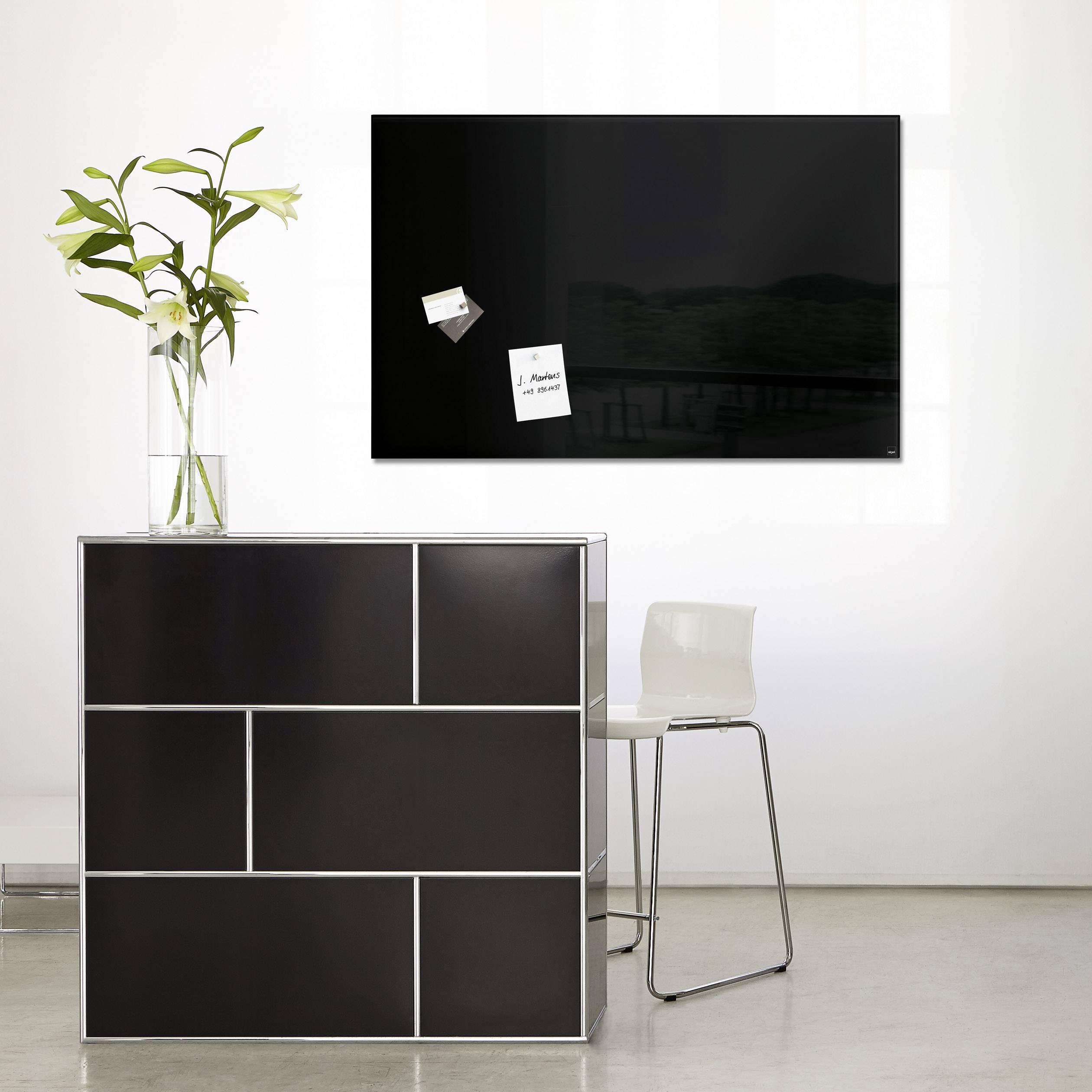 Un espace moderne avec un tableau noir accroché au mur, en dessous duquel se trouve une table minimaliste noire aux bords blancs, et deux fleurs dans un vase.