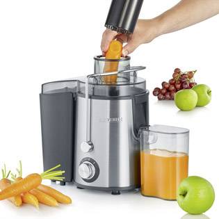 Une main presse une carotte dans un extracteur de jus. Une cruche recueille le jus. À côté, il y a des carottes, des pommes vertes et des raisins.