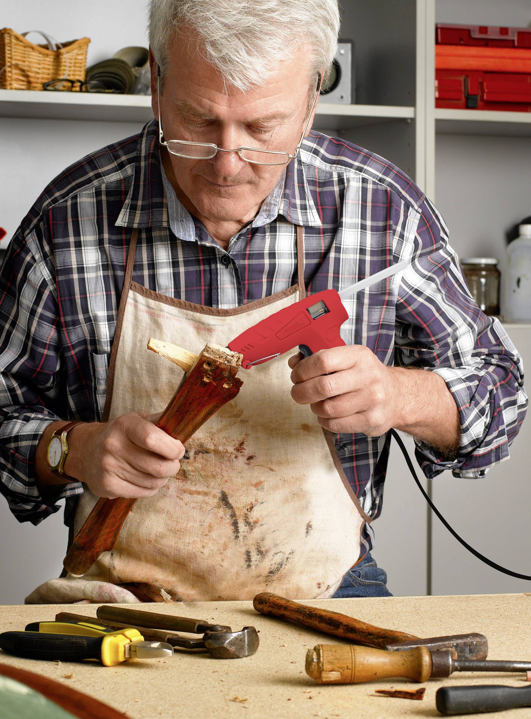 Un homme âgé répare un pied de chaise avec un pistolet à colle chaude dans un atelier, entouré d'outils sur une table.