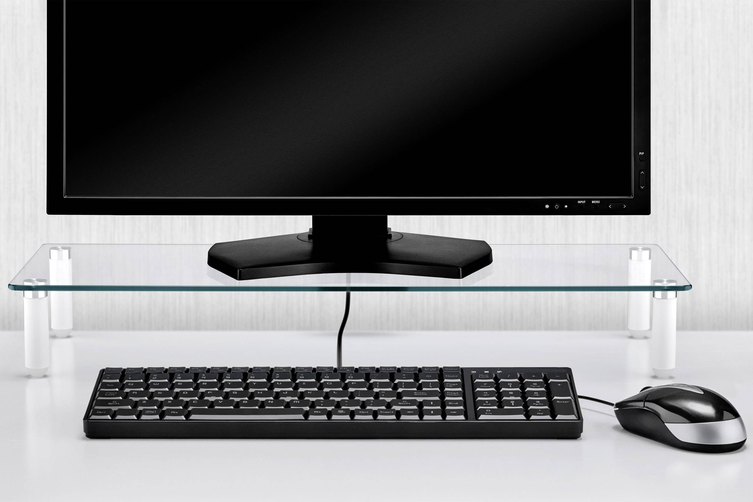 Un ordinateur avec écran posé sur un support en verre, avec un clavier et une souris en dessous sur une table blanche.