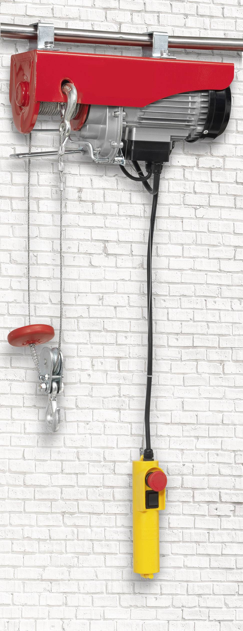 Treuil électrique à boîtier rouge monté sur un mur de briques blanches. Une télécommande jaune est suspendue au câble.