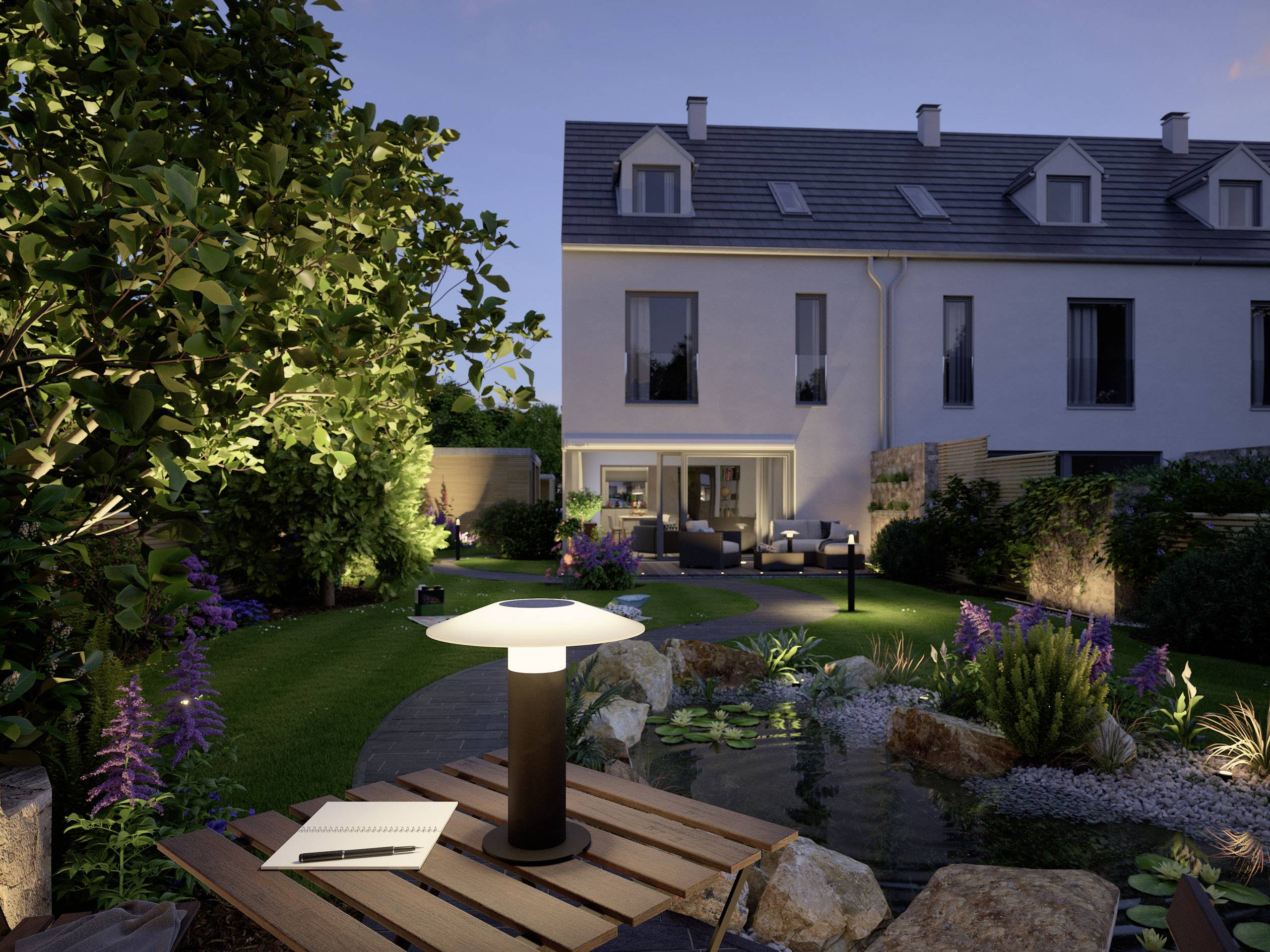 Jardin à la lumière du soir avec une table en pierre éclairée, un cahier et une lampe au premier plan ; maison moderne et mobilier de jardin en arrière-plan.