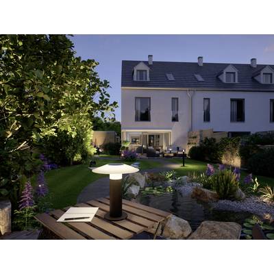 Jardin à la lumière du soir avec une table en pierre éclairée, un cahier et une lampe au premier plan ; maison moderne et mobilier de jardin en arrière-plan.