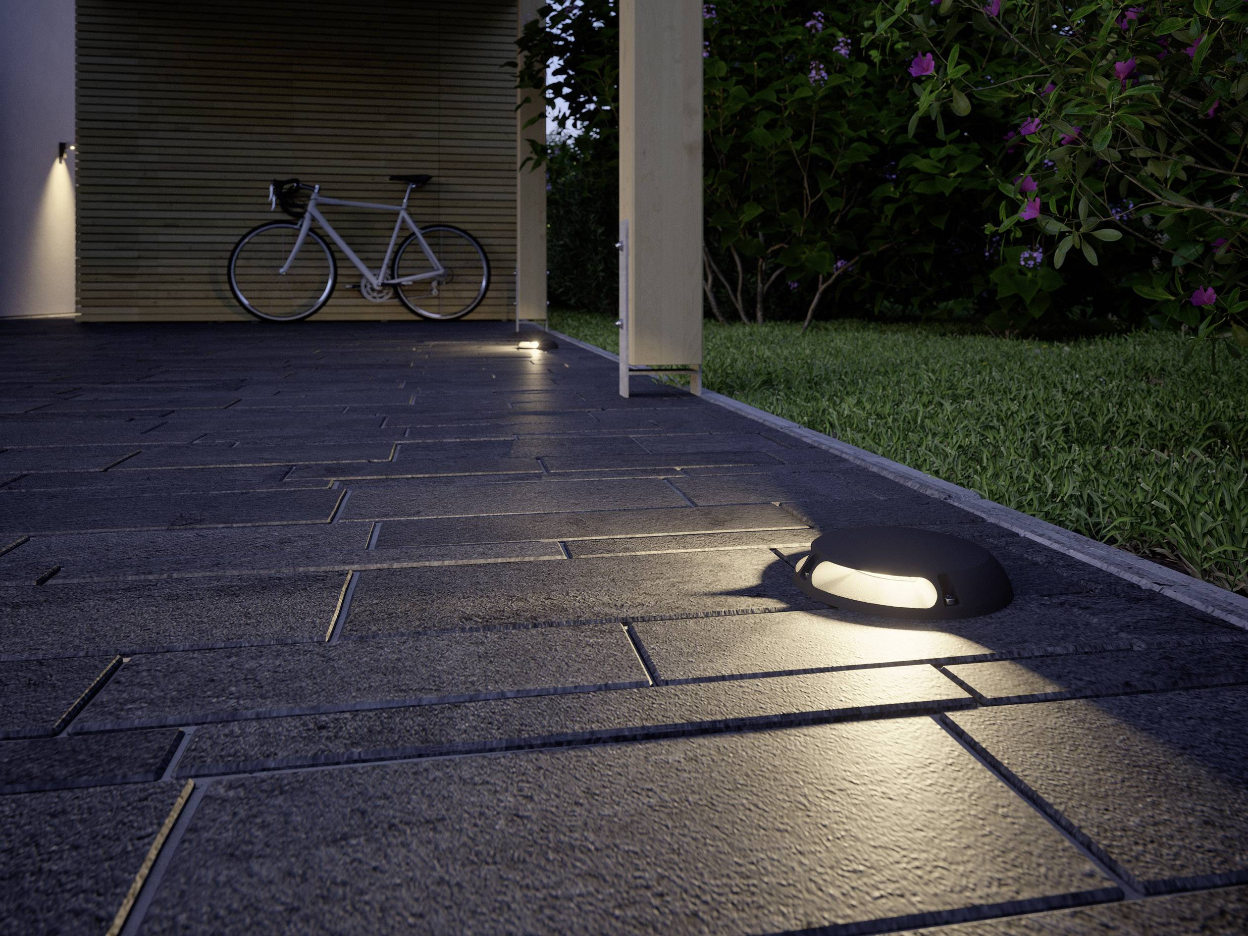 Allée pavée la nuit, éclairée par des luminaires au sol ; un vélo contre un mur en bois, des plantes à droite en arrière-plan.