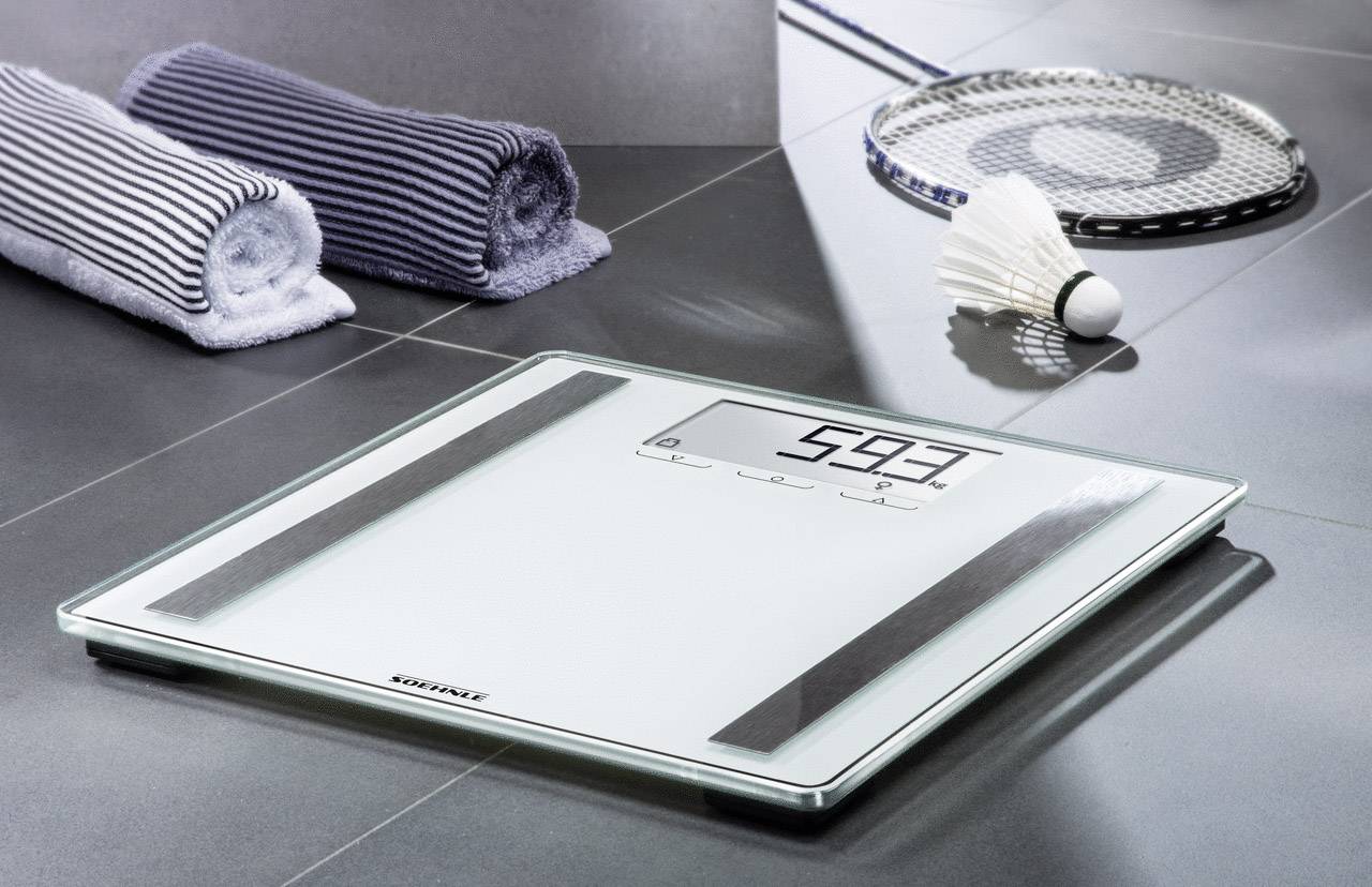 Balance numérique posée sur un carrelage de salle de bain, affichant 59,3 kg. À côté, deux serviettes pliées et une raquette de badminton avec un volant.