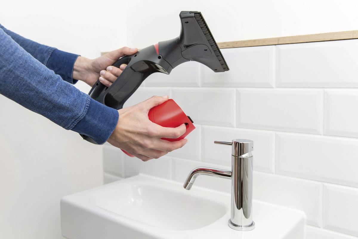 Une personne tient un nettoyeur de vitres électrique au-dessus d'un lavabo dans une salle de bain aux carreaux blancs.
