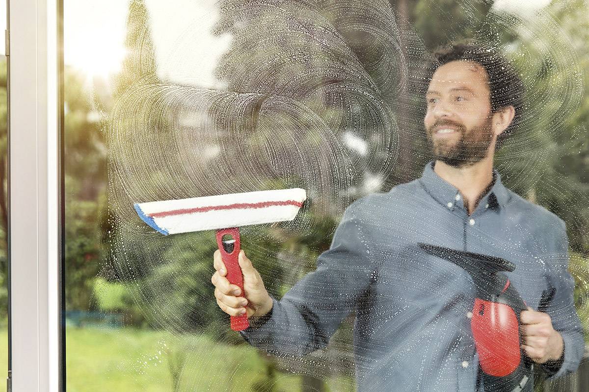 Un homme nettoie une fenêtre avec un aspirateur à vitres devant un arrière-plan flou. La lumière du soleil traverse la fenêtre.