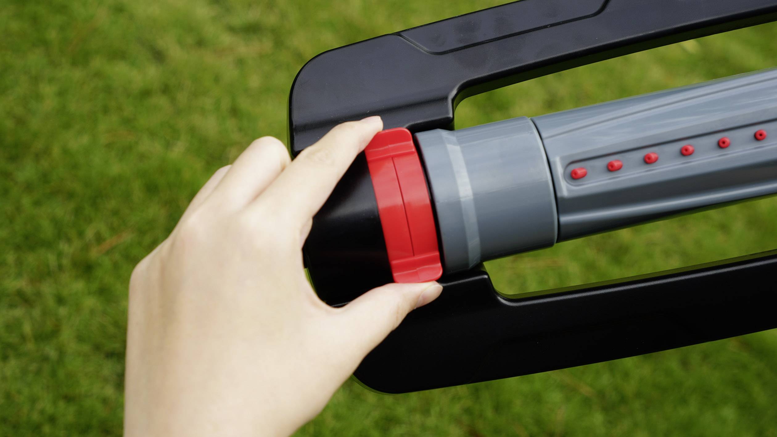 Une main tourne un bouton rouge sur un appareil de jardin devant un fond d'herbe verte.